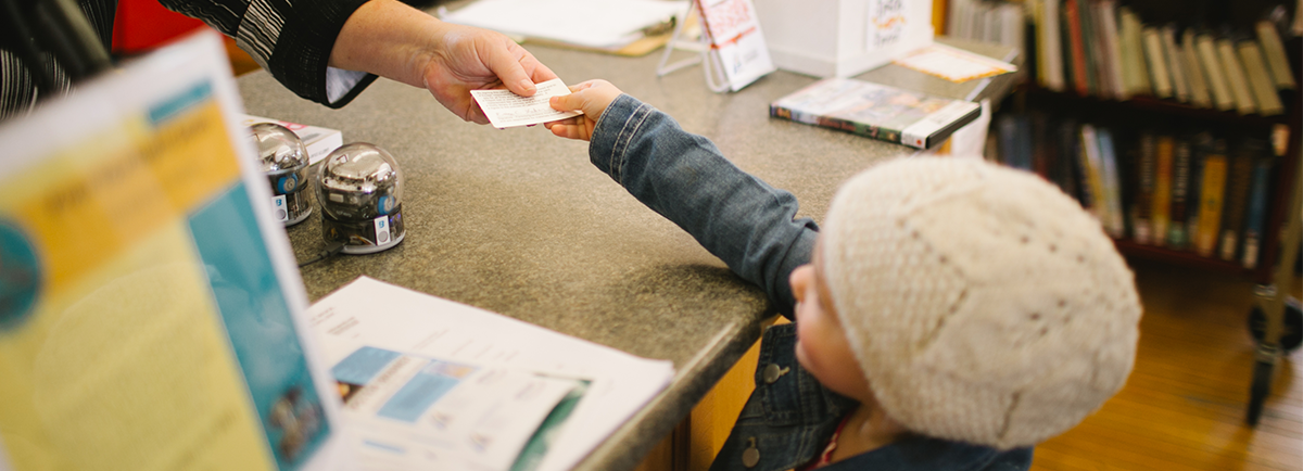 A child handing her library card to a staff member at the service desk.