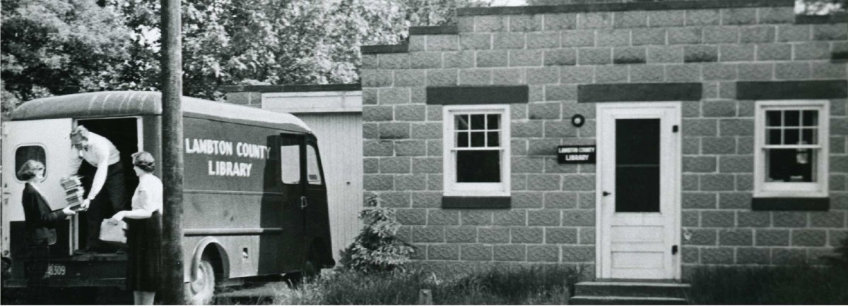 Women unloading books from a van next to a Lambton County Library building. 