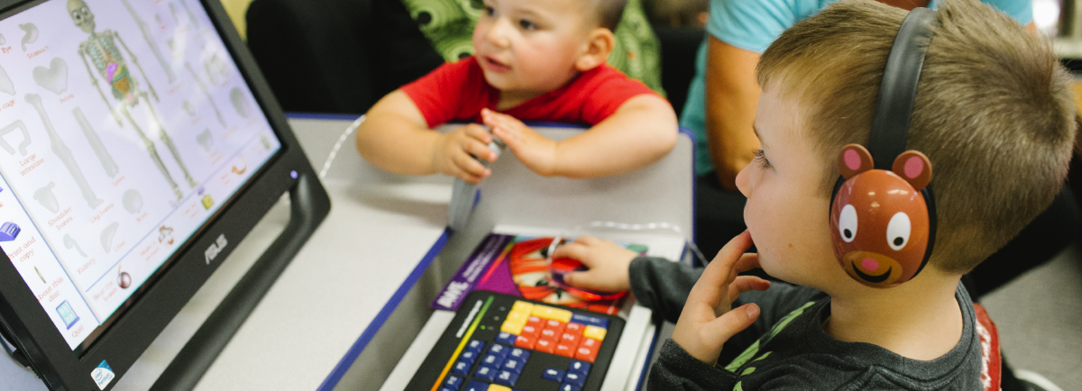 Kid at the computer while a younger kid looks on.