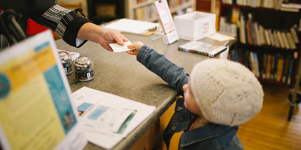 Child handing library card to librarian across a desk.