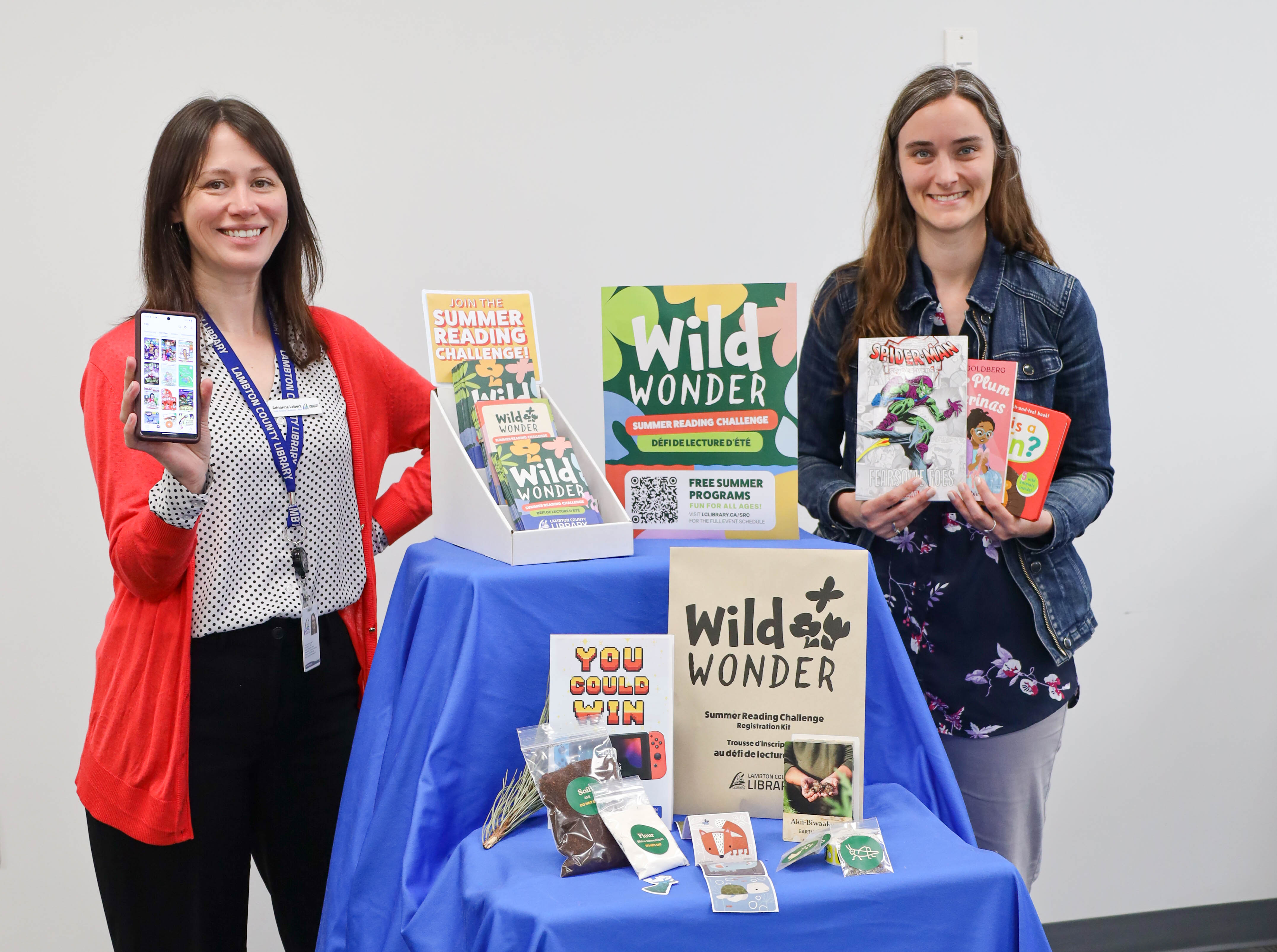 Library staff pose with the 2025 Summer Reading Challenge information, book prizes, and a phone with the reading list app on screen.