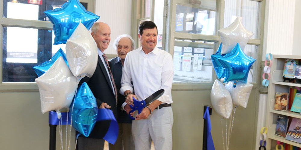 Three men cutting a blue ribbon in a library.