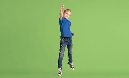 Kid with blond hair jumping and giving a thumbs up in front of a green background.
