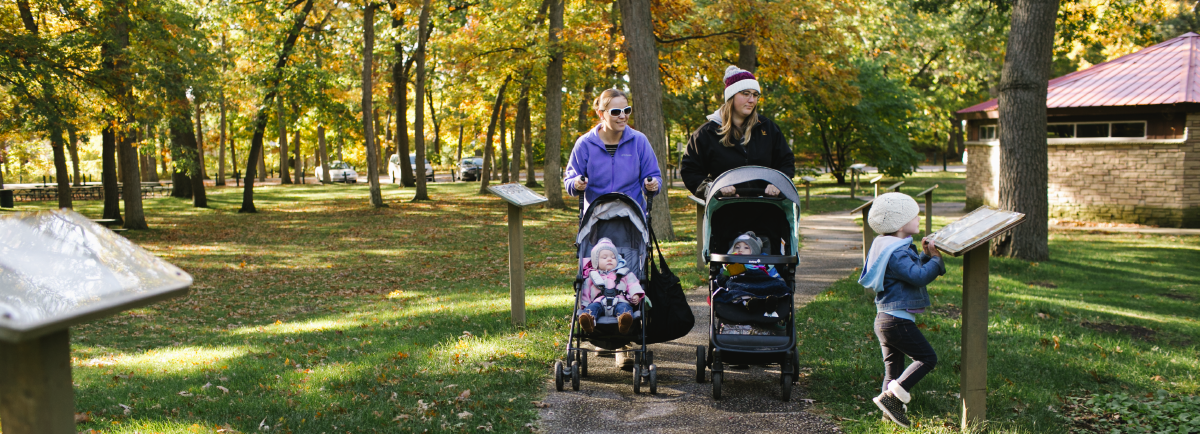 Two people with strollers and a young girl walking in the park.