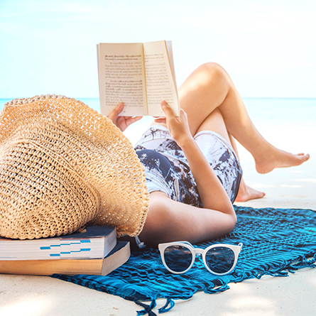 Person laying in sand on back holding up a book and reading.