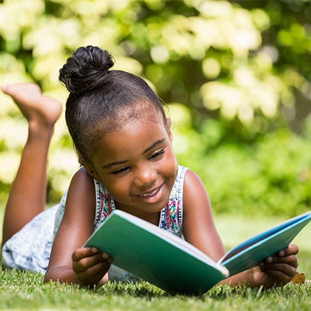 Black girl laying in grass outside reading a book.