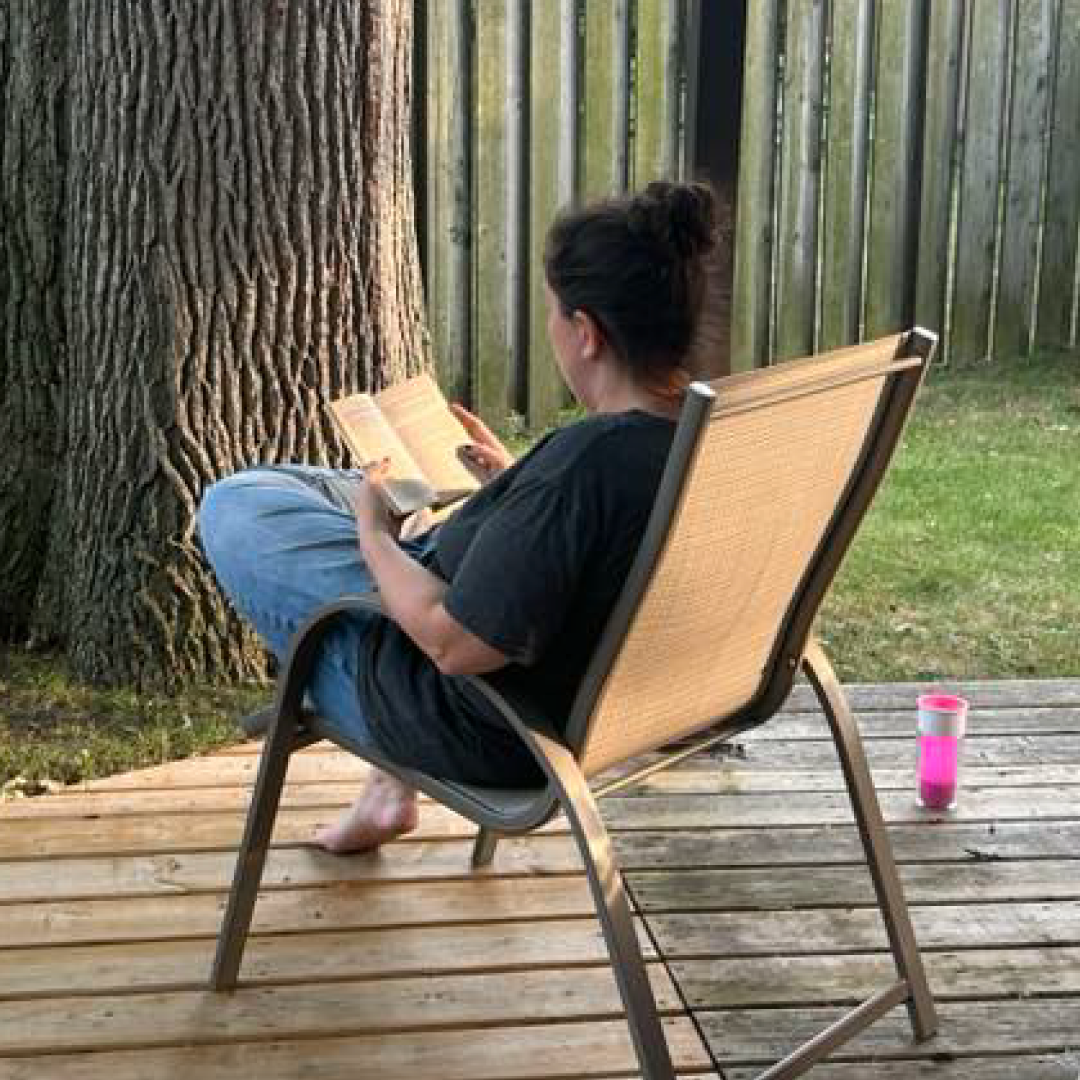 Person sitting in a chair on a wooden deck, reading a book beside a large tree.