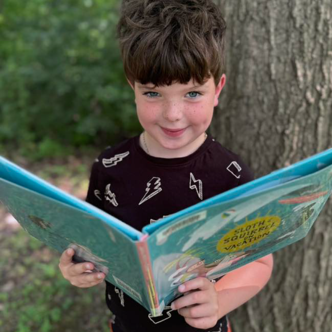 Child standing outdoors, holding and reading an open book near a tree.
