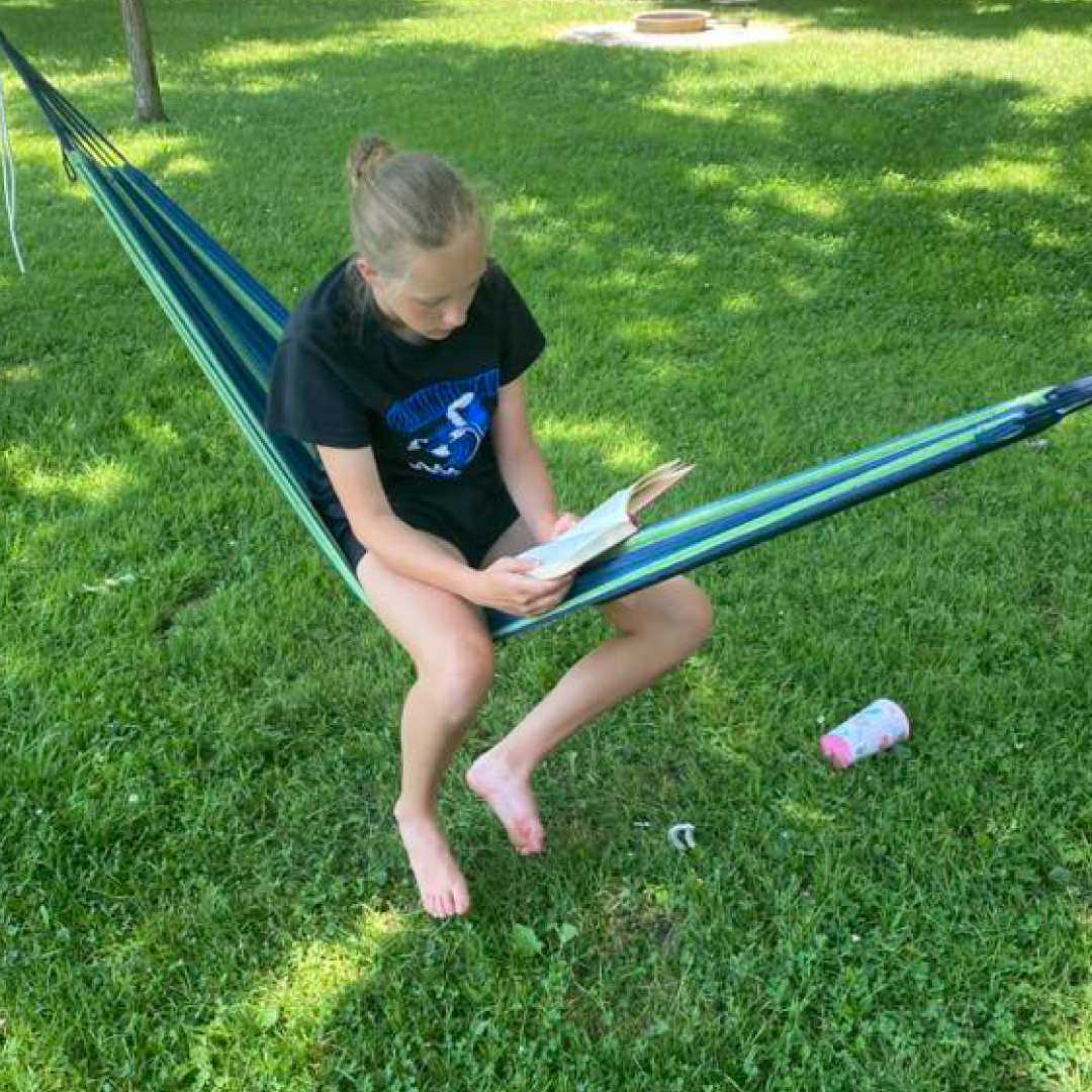 Person sitting in a hammock on grass, reading a book in a backyard setting.