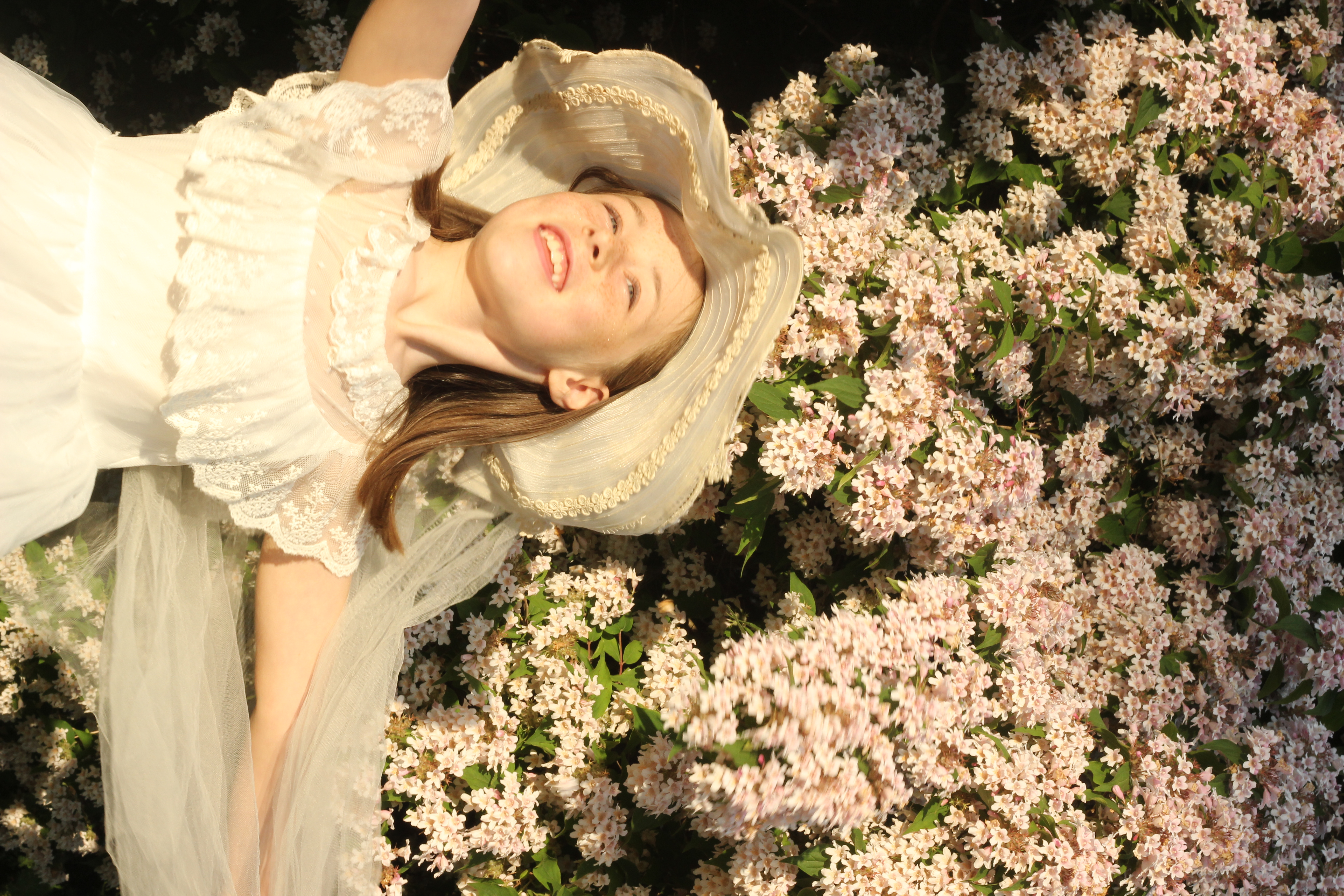 A young girl, dressed in a white gown and hat, poses whimsically in front of a wall of flowers