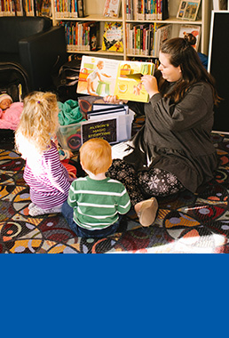 Lady holding out a book and reading to two children sitting on a carpet.