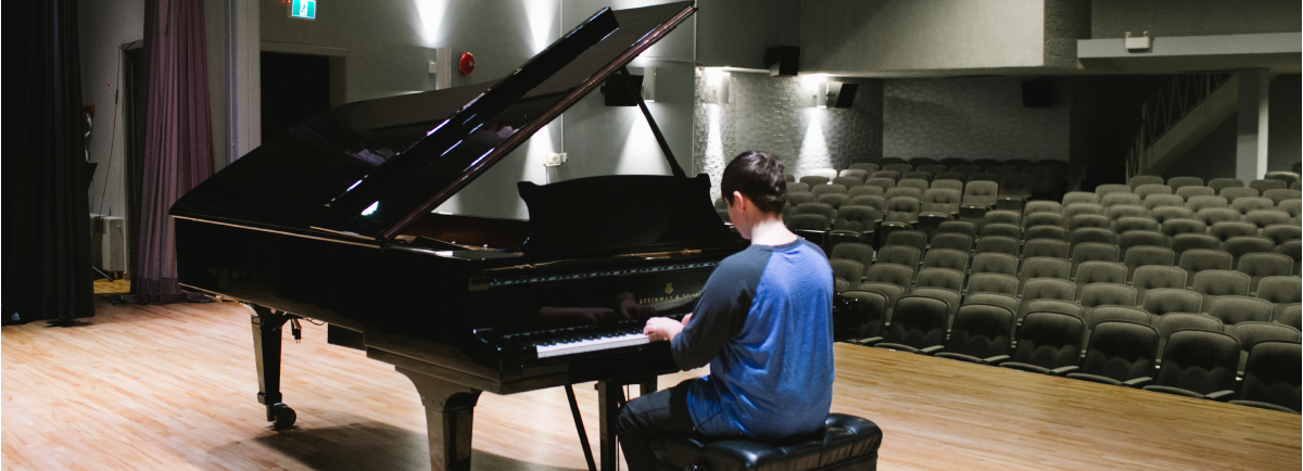 Person sitting at a piano on a stage with seating in the background. 